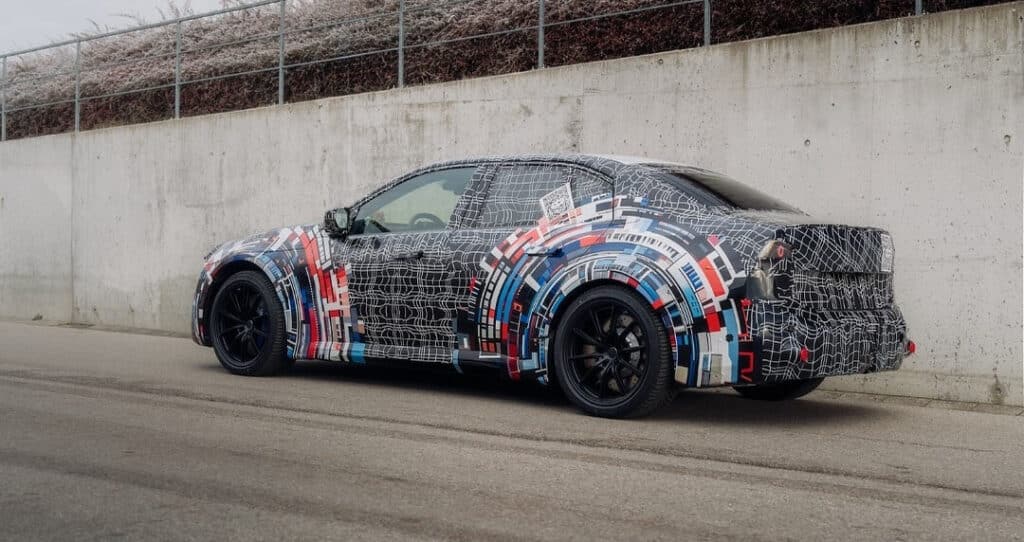 BMW M3 eléctrico estacionado junto a un muro de hormigón gris. El camuflaje del vehículo combina un patrón de rejilla digital en blanco y negro con círculos concéntricos en rojo y azul alrededor de las ruedas, destacando sus líneas deportivas y musculosas