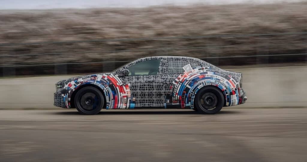 Prototipo de BMW M3 eléctrico fotografiado en movimiento, cubierto con un llamativo camuflaje que mezcla patrones geométricos en blanco y negro con acentos en rojo y azul alrededor de las ruedas. El coche mantiene la silueta deportiva característica del M3 y monta llantas negras deportivas