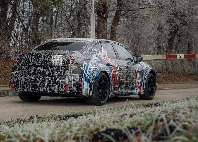 Vista trasera del BMW M3 eléctrico en un entorno boscoso invernal. El camuflaje permite distinguir los pilotos LED traseros, el difusor deportivo y la línea del maletero. Las llantas negras y la postura ancha enfatizan su carácter deportivo