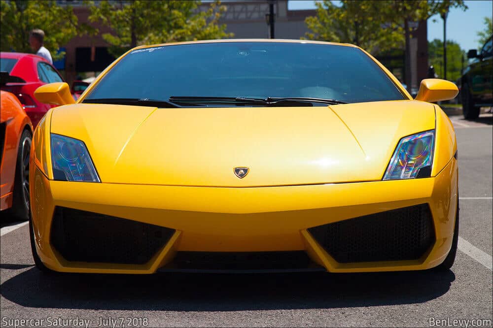 Imagen de un automóvil Lamborghini Gallardo de color amarillo estacionado en un evento. El coche está posicionado de frente y en el centro, mostrando sus distintivos faros angulares y su diseño elegante. El evento parece ser 'Supercar Saturday', celebrado el 7 de julio de 2018, según indica el texto en la esquina inferior izquierda de la imagen. En la esquina inferior derecha se menciona el sitio web 'BenLevy.com'.