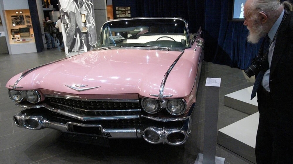 Cadillac clásico de color rosa expuesto en un museo, con un hombre mayor observándolo de cerca.