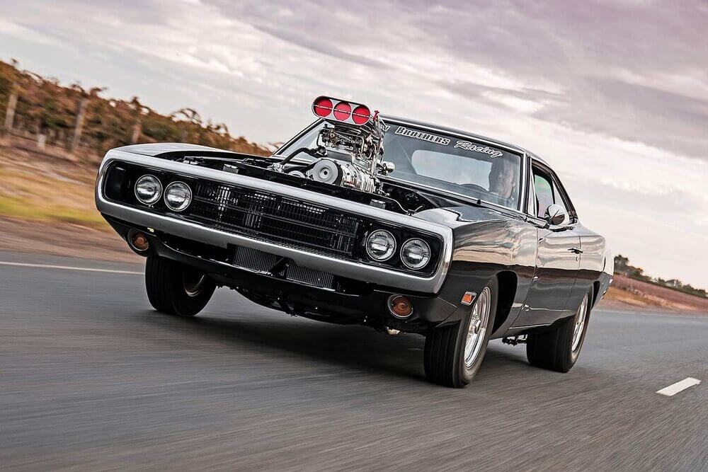 La imagen muestra un coche clásico de músculo, específicamente un Dodge Charger negro, conduciendo por una carretera. El coche está equipado con un gran supercargador expuesto que sobresale del capó, una característica notable de los vehículos de alto rendimiento. La parte delantera del coche está ligeramente levantada, lo que sugiere que está acelerando rápidamente. El parabrisas tiene una calcomanía que dice "BROTHERS Racing". El fondo muestra un paisaje borroso, indicando que el coche está en movimiento.