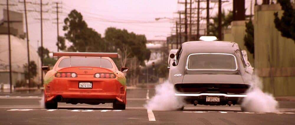 La imagen muestra una escena de la película "Fast and Furious 1" donde dos coches están en una carrera callejera. A la izquierda, hay un Toyota Supra naranja con un alerón grande y detalles de pintura verde. A la derecha, hay un Dodge Charger negro con humo saliendo de las llantas traseras, indicando que está acelerando rápidamente. Ambos coches están en una calle urbana con líneas eléctricas y árboles en el fondo.