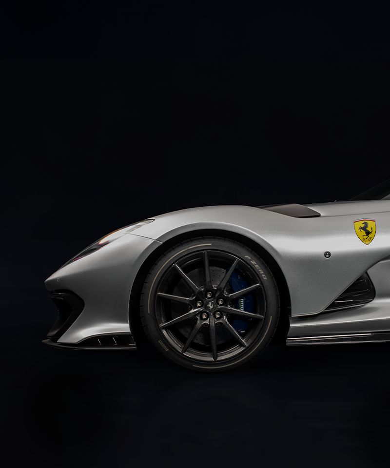 La imagen muestra el lado delantero de un Ferrari Competizione A plateado, parte del programa 'Ferrari Tailor Made'. El enfoque está en la rueda delantera y el guardabarros, destacando el diseño intrincado de la llanta de aleación negra con pinzas de freno azules, las líneas aerodinámicas elegantes del coche y el icónico logotipo de Ferrari en el guardabarros. El fondo oscuro enfatiza la apariencia elegante y deportiva del coche.