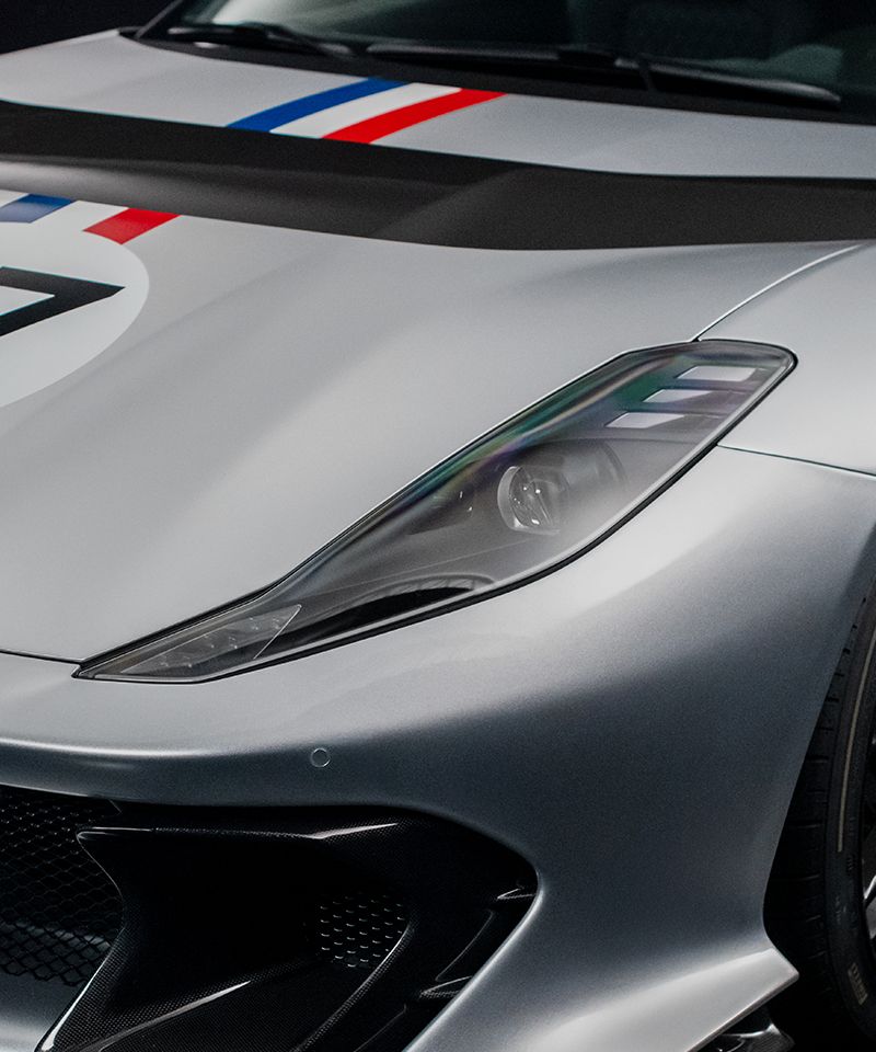 "La imagen muestra un primer plano del frente de un Ferrari Competizione A, parte del programa 'Ferrari Tailor Made'. Se puede observar el faro delantero derecho del automóvil, que tiene un diseño estilizado y moderno. El capó del coche presenta una franja negra y dos franjas más pequeñas, una azul y otra roja, que son características de la personalización 'Tailor Made'. También se aprecia parte de una gran calcomanía circular con el número 7 en el capó. La imagen destaca por la elegancia y el diseño exclusivo del vehículo, resaltando la atención al detalle y la personalización que ofrece Ferrari a sus clientes.