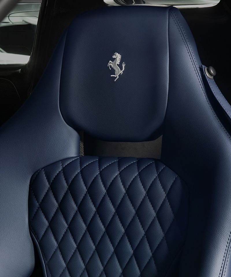 La imagen muestra el asiento de un automóvil Ferrari, específicamente un Ferrari Competizione A, que forma parte del programa 'Ferrari Tailor Made'. El asiento está tapizado en cuero azul con un patrón de costura en forma de diamante en la parte inferior. En la parte superior del respaldo del asiento, se encuentra el logotipo de Ferrari, un caballo encabritado, bordado en color plateado. Esta imagen es interesante y relevante porque destaca la atención al detalle y la personalización que ofrece Ferrari a sus clientes a través de su programa de personalización 'Tailor Made'.