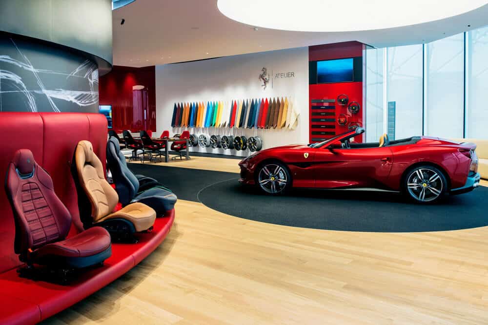 "La imagen muestra un interior de un showroom de Ferrari, específicamente el área de 'Ferrari Tailor Made'. En el centro de la imagen hay un Ferrari descapotable rojo. A la izquierda, hay una fila de asientos de coche de diferentes colores y estilos, montados en una estructura roja. En la pared del fondo, se pueden ver muestras de colores y materiales para personalizar los vehículos, así como varias llantas y volantes. La imagen es interesante y relevante porque muestra las opciones de personalización exclusivas que ofrece Ferrari a sus clientes, permitiéndoles diseñar un coche a medida según sus preferencias.