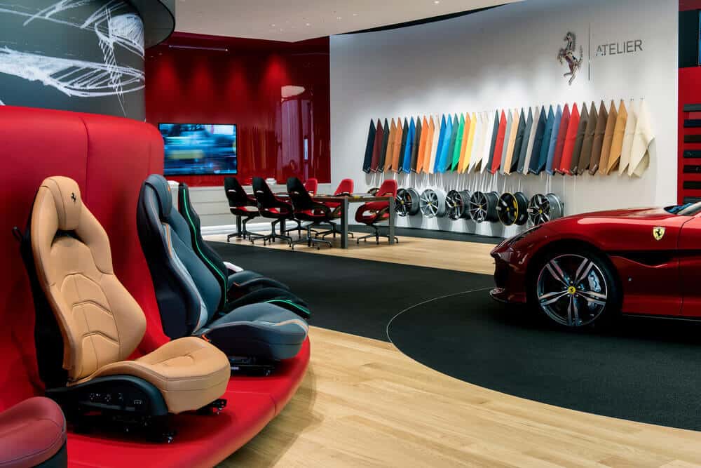 "La imagen muestra un lujoso showroom de Ferrari Tailor Made. En el centro de la imagen hay un Ferrari descapotable rojo. A la izquierda, hay una fila de asientos de coche de diferentes colores y diseños, montados en una estructura curva roja. En la pared del fondo, se exhiben múltiples opciones de colores y materiales para personalizar los vehículos, junto con varios volantes y llantas. El ambiente es moderno y elegante, con una combinación de suelos de madera clara y alfombras negras. La imagen es interesante porque destaca las opciones de personalización exclusivas que ofrece Ferrari a sus clientes, permitiéndoles crear un coche único y a medida.