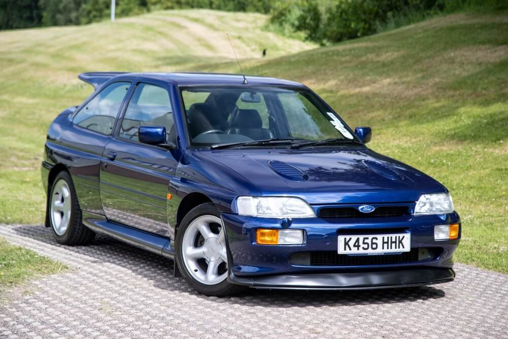 La imagen muestra un Ford Escort Cosworth azul de 1992 estacionado en una zona pavimentada con un entorno de césped. El vehículo presenta un distintivo alerón trasero, llantas de aleación y una matrícula delantera con el número de registro "K456 HHK". Este modelo es notable por su rendimiento y su herencia en el mundo de los rallies, lo que lo convierte en un ejemplo interesante y relevante de los coches deportivos de los años 90.