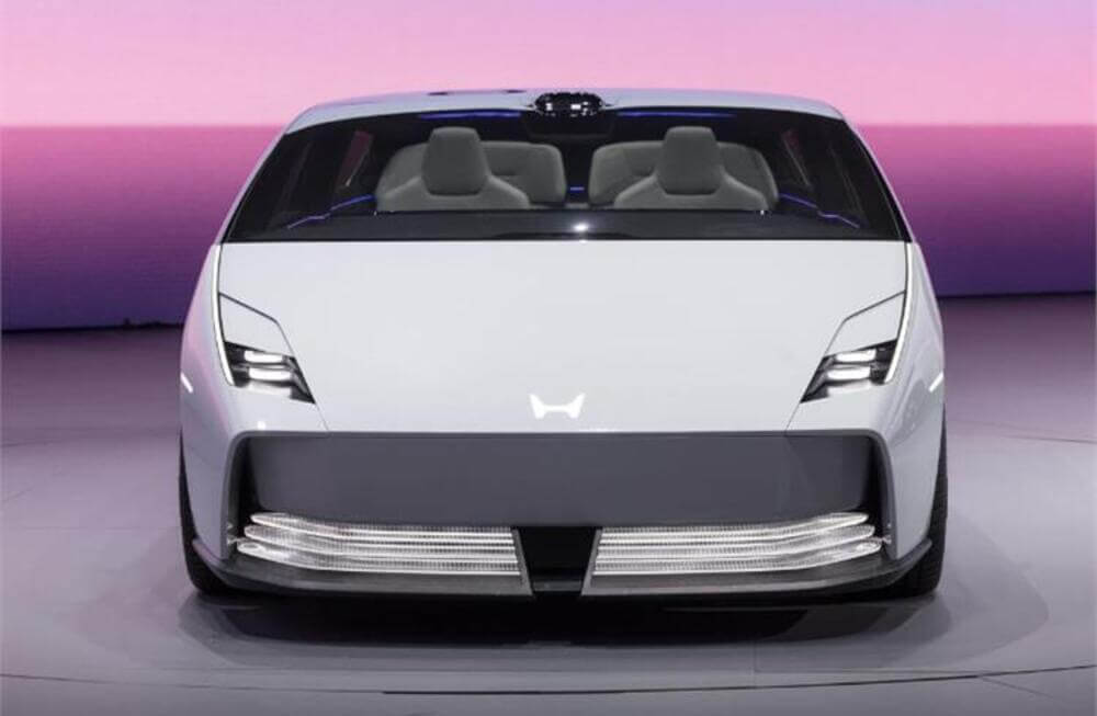 Imagen de un automóvil Honda 0 Saloon de color blanco visto desde el frente. El coche tiene una forma aerodinámica y suave, con faros angulares y una parrilla frontal distintiva. El interior es visible a través del parabrisas, mostrando dos asientos. El vehículo parece ser un concepto o prototipo, probablemente mostrando características avanzadas de diseño y tecnología.