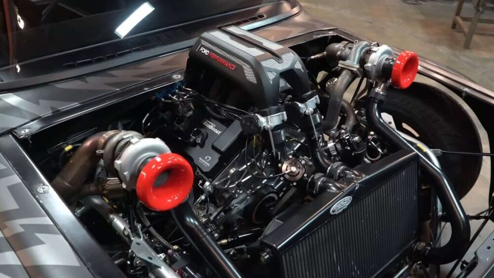 La imagen muestra el compartimento del motor del Hoonitruck. El motor es un Ford Performance EcoBoost V6, que se destaca con la marca "Ford Performance" en la cubierta del motor. La configuración del motor incluye dos grandes turbocompresores con entradas rojas, lo que indica un sistema de alto rendimiento y doble turbo. La presencia de estos turbocompresores sugiere que el motor está diseñado para una potencia significativa, lo que resulta interesante y relevante para los entusiastas del automovilismo y aquellos interesados en vehículos de alto rendimiento. La configuración detallada y limpia del compartimento del motor resalta la ingeniería y personalización involucradas en esta construcción.