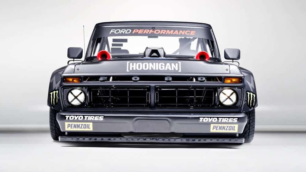 La imagen muestra una vista frontal de una camioneta Ford modificada conocida como el "Hoonitruck". La camioneta presenta una marca prominente y modificaciones, incluyendo:

La palabra "FORD" en la parrilla frontal.

La palabra "[HOONIGAN]" en el parabrisas.

"FORD PERFORMANCE" escrito en la parte superior del parabrisas.

Logotipos de "TOYO TIRES" y "PENNZOIL" en el parachoques delantero.

Logotipos de Monster Energy en los lados del parachoques delantero.

Grandes entradas de aire rojas en el capó.

Una apariencia robusta y lista para la carrera con luces adicionales y un parachoques delantero reforzado.

Esta imagen es interesante y relevante porque muestra un vehículo altamente personalizado y orientado al rendimiento, probablemente utilizado para deportes de motor o eventos promocionales. La marca y las modificaciones sugieren una colaboración entre varias marcas automotrices y de estilo de vida.