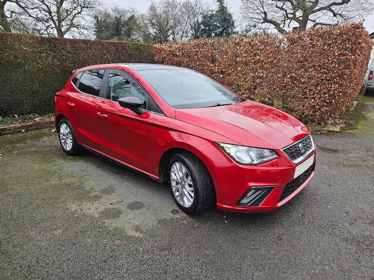 La imagen muestra un coche hatchback rojo estacionado en una entrada pavimentada. El coche está limpio y parece estar en buen estado. El fondo incluye un seto con hojas marrones y algunos árboles, lo que indica que podría ser otoño o invierno. Esta imagen es ficticia y se utiliza para un estudio que demuestra las habilidades de marketing de Miana.
