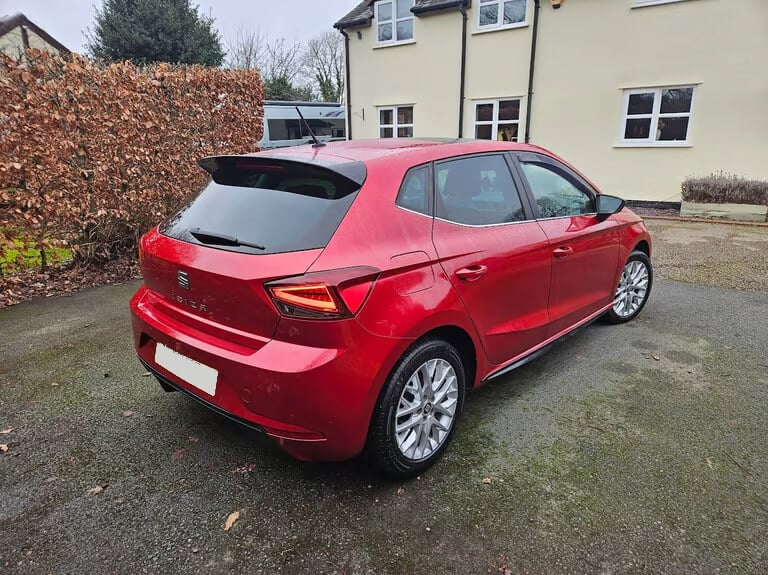 La imagen muestra un coche SEAT Ibiza rojo estacionado en una entrada pavimentada frente a una casa. El coche está posicionado en un ángulo, mostrando la parte trasera y el lado derecho. La casa en el fondo es de color crema con ventanas enmarcadas en blanco. Hay un seto a la izquierda de la imagen y la entrada parece estar mojada, lo que sugiere lluvia reciente. La matrícula del coche está oculta por privacidad. Esta imagen es ficticia y se utiliza para un estudio que demuestra las habilidades de marketing de Miana.