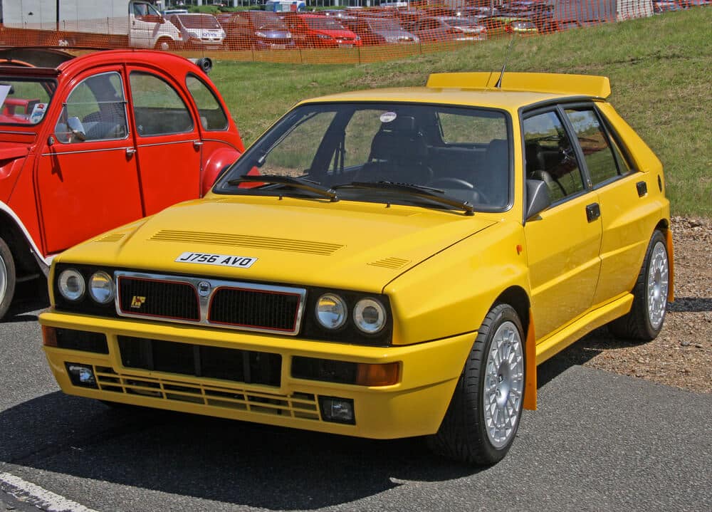 La imagen muestra un Lancia Delta HF Integrale amarillo estacionado junto a un Citroën 2CV rojo. El Lancia Delta HF Integrale es una versión de alto rendimiento del Delta, conocida por su éxito en las carreras de rally a finales de los años 80 y principios de los 90. El coche presenta un diseño distintivo y cuadrado con un prominente alerón trasero, grandes llantas de aleación y una parrilla delantera con el emblema de Lancia. La matrícula dice "J756 AVO". El Citroën 2CV en el fondo es un coche económico clásico francés, que contrasta con la naturaleza deportiva del Lancia.