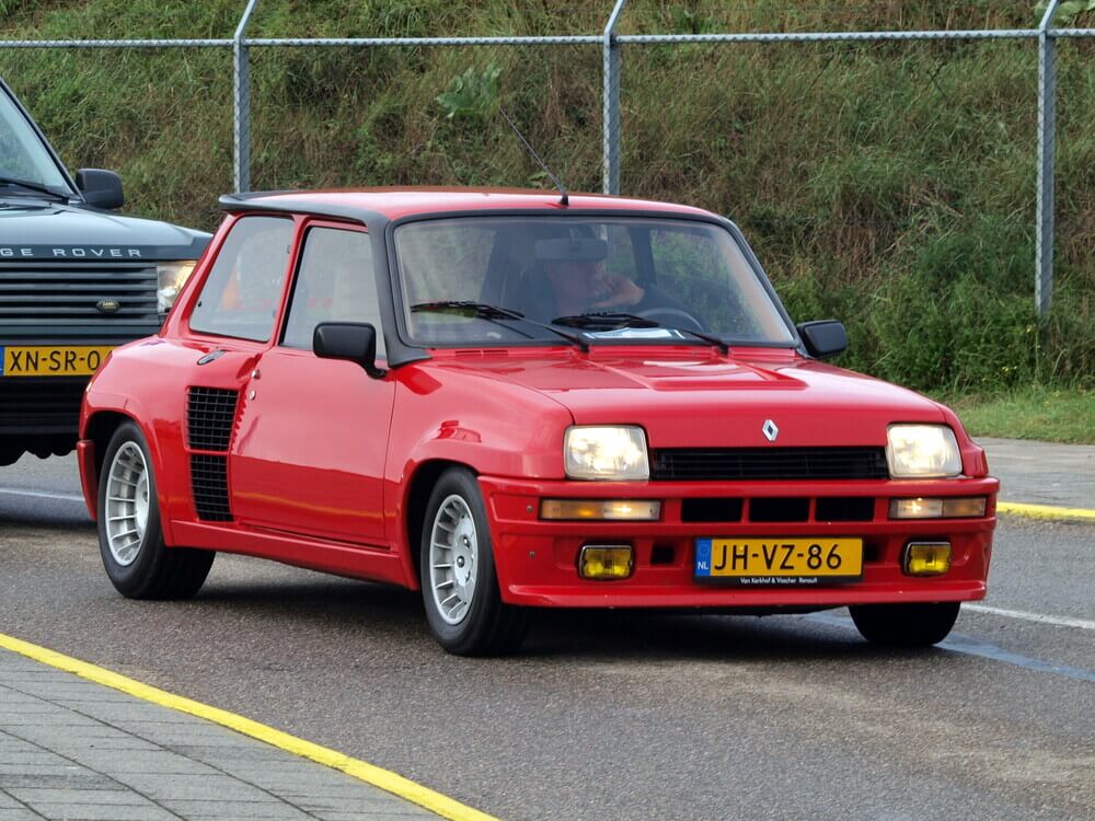 La imagen muestra un Renault 5 Turbo rojo, un hatchback de alto rendimiento, conduciendo por una carretera. El coche se destaca por su diseño cuadrado distintivo, carrocería ancha y disposición de motor central, lo que lo diferencia del Renault 5 estándar. La matrícula del coche dice "JH-VZ-86" y tiene una placa amarilla de la Unión Europea con una franja azul en el lado izquierdo que indica que está registrado en los Países Bajos. En el fondo, hay un Range Rover verde con una matrícula "XN-SR-04" también de los Países Bajos.