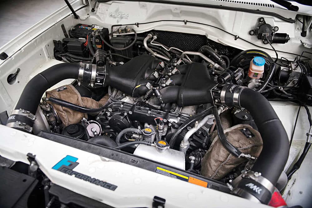 "El compartimento del motor de un Nissan Patrol Y61 modificado. El motor ha sido ampliamente modificado, presentando un sistema de doble turbo con grandes tuberías de intercooler y componentes de posventa. Las modificaciones sugieren un aumento significativo en el rendimiento y la potencia en comparación con el motor de serie. La presencia de piezas de alto rendimiento, como los turbocompresores, tuberías personalizadas y colectores de escape envueltos en material aislante, indica que este vehículo probablemente se utiliza para aplicaciones de alto rendimiento, posiblemente en escenarios todoterreno o de carreras. La marca 'F Performance' en la parte frontal del compartimento del motor enfatiza aún más la naturaleza orientada al rendimiento de las modificaciones.