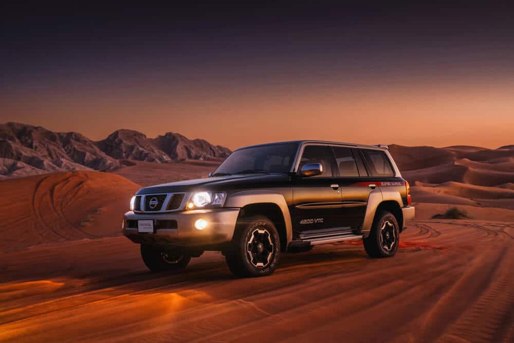 Un Nissan Patrol Y61 SUV está estacionado en un paisaje desértico arenoso durante el atardecer o amanecer. El vehículo está en primer plano, con los faros encendidos, iluminando la arena. El fondo presenta dunas de arena onduladas y montañas escarpadas, creando un entorno dramático y aventurero. La iluminación y la composición destacan las capacidades robustas y todoterreno del vehículo, haciendo que sea una representación interesante y relevante para mostrar el rendimiento del Nissan Patrol Y61 en terrenos desafiantes. El texto 'PATROL' y '4800 VTC' es visible en el vehículo, indicando su modelo y tipo de motor.