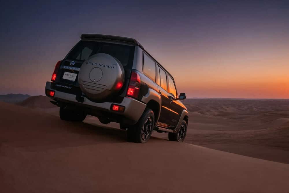 Un Nissan Patrol Super Safari SUV está conduciendo sobre dunas de arena al atardecer. El vehículo está posicionado en un ángulo, destacando sus capacidades todoterreno. El fondo presenta un vasto paisaje desértico con un cielo colorido que pasa de tonos naranjas a púrpuras, indicando la puesta del sol. Esta imagen es interesante y relevante ya que muestra el diseño robusto del vehículo y su idoneidad para terrenos desérticos, enfatizando su naturaleza aventurera y duradera.