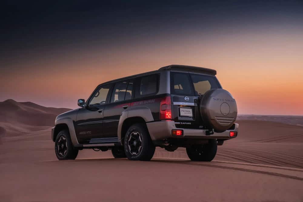 "Un Nissan Patrol Y61, también conocido como Super Safari, está estacionado en un paisaje desértico durante el atardecer. El vehículo está posicionado con la parte trasera hacia la cámara, mostrando su construcción robusta y sus capacidades todoterreno. El fondo presenta dunas de arena y un cielo en degradado que pasa de azul profundo a tonos cálidos de naranja, indicando el amanecer o el atardecer. Los detalles de la marca y el modelo del vehículo, como 'Super Safari' y '4800 VTC', son visibles, enfatizando su potente motor y su idoneidad para terrenos difíciles. Esta imagen es interesante y relevante ya que destaca el diseño y la funcionalidad del Patrol Y61 en un entorno donde dicho vehículo sería más útil.