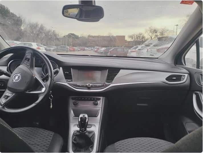 "La imagen muestra el interior de un Opel Astra de segunda mano en Zaragoza. Se pueden ver los asientos delanteros, el salpicadero y el volante. El coche está estacionado en un aparcamiento con otros vehículos visibles a través del parabrisas. El salpicadero cuenta con una pantalla central, rejillas de ventilación y varios controles. El volante tiene el logotipo de Opel en el centro y hay una palanca de cambios en la consola central. Los asientos tienen un tejido con patrón. Esta imagen es ficticia y se utiliza para un estudio que demuestra las habilidades de marketing de Miana.