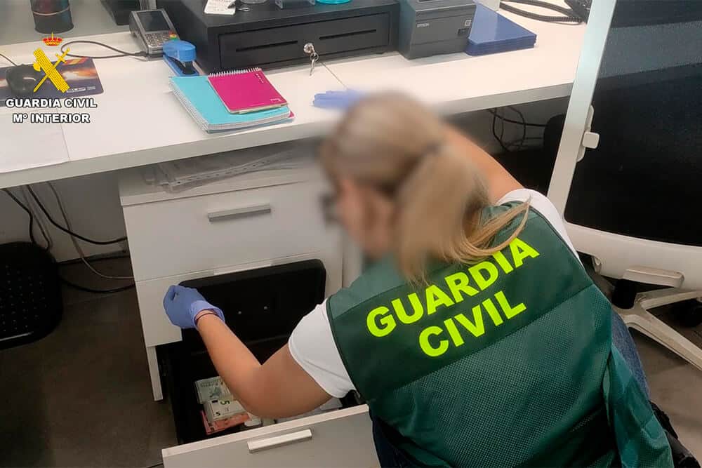 La imagen muestra a un miembro de la Guardia Civil, identificado por el chaleco verde con letras amarillas, inspeccionando un cajón en un entorno de oficina. La persona lleva guantes azules y parece estar examinando el contenido de una caja fuerte o cajón, que incluye varios objetos como dinero en efectivo. La escena sugiere una investigación oficial o registro realizado por la Guardia Civil. El logo de la Guardia Civil y el texto "GUARDIA CIVIL M° INTERIOR" son visibles en la esquina superior izquierda de la imagen.