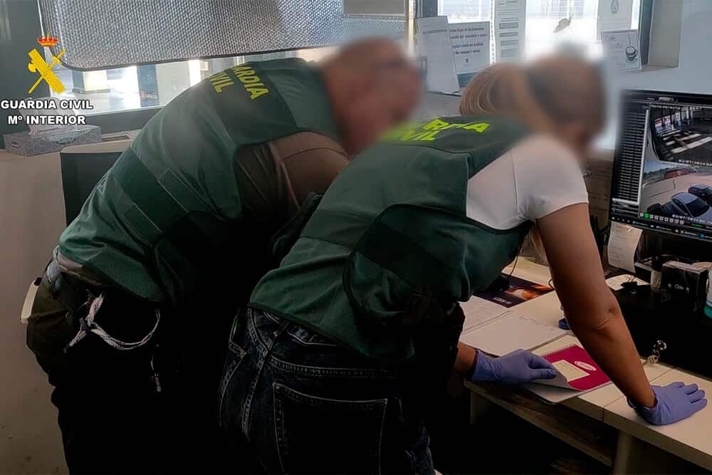 La imagen muestra a dos personas con chalecos verdes con el texto "GUARDIA CIVIL" en la espalda. Están inclinados sobre un escritorio, examinando documentos y una pantalla de computadora. Una de las personas lleva guantes azules y sostiene un pasaporte rosa. La pantalla de la computadora muestra imágenes de vigilancia de vehículos. El entorno parece ser una oficina o sala de control, probablemente relacionada con actividades de control fronterizo o aplicación de la ley. La imagen es interesante y relevante, ya que muestra el trabajo de la Guardia Civil en la supervisión e inspección de documentos e imágenes de vigilancia.