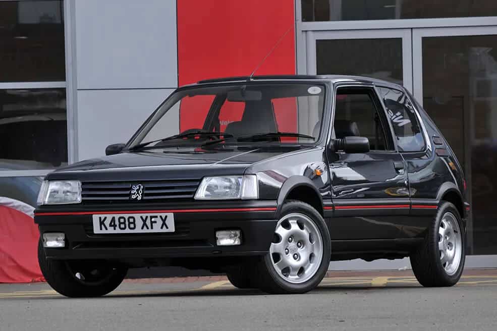 La imagen muestra un Peugeot 205 GTI negro estacionado frente a un edificio. El coche tiene un diseño distintivo con una franja roja a lo largo del lateral, llantas de aleación y una matrícula delantera que dice "K488 XFX". El logotipo de Peugeot se muestra prominentemente en la parrilla delantera.
