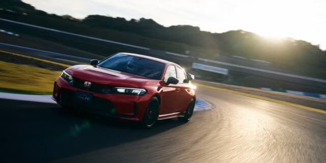 Honda Civic Type R 2025 en color rojo intenso fotografiado en movimiento dinámico en una pista de carreras durante el atardecer. El auto está tomando una curva, mostrando su perfil deportivo con detalles en negro, mientras el sol poniente crea un efecto dramático de iluminación sobre la carrocería.