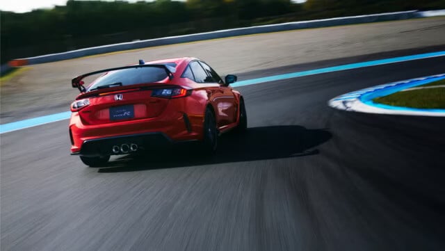 Vista trasera del Honda Civic Type R 2025 en color rojo tomando una curva en una pista de carreras. Se aprecia el movimiento dinámico del vehículo, con los tres tubos de escape centrales característicos y el alerón deportivo negro. Los bordes de la pista están marcados con líneas azules y blancas, y la imagen captura el efecto de velocidad con un ligero desenfoque de movimiento.