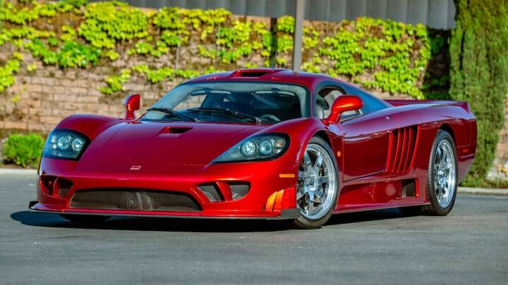 La imagen muestra un automóvil deportivo de alta gama, específicamente un Saleen S7, de color rojo brillante. El coche está estacionado en una superficie pavimentada con un fondo de vegetación verde y una pared de ladrillos. El diseño del Saleen S7 es aerodinámico y agresivo, con faros delanteros de forma ovalada, una parrilla baja y ancha, y llantas de aleación cromadas. Este vehículo es conocido por su alto rendimiento y diseño exclusivo, lo que lo hace interesante y relevante para los entusiastas de los automóviles deportivos.