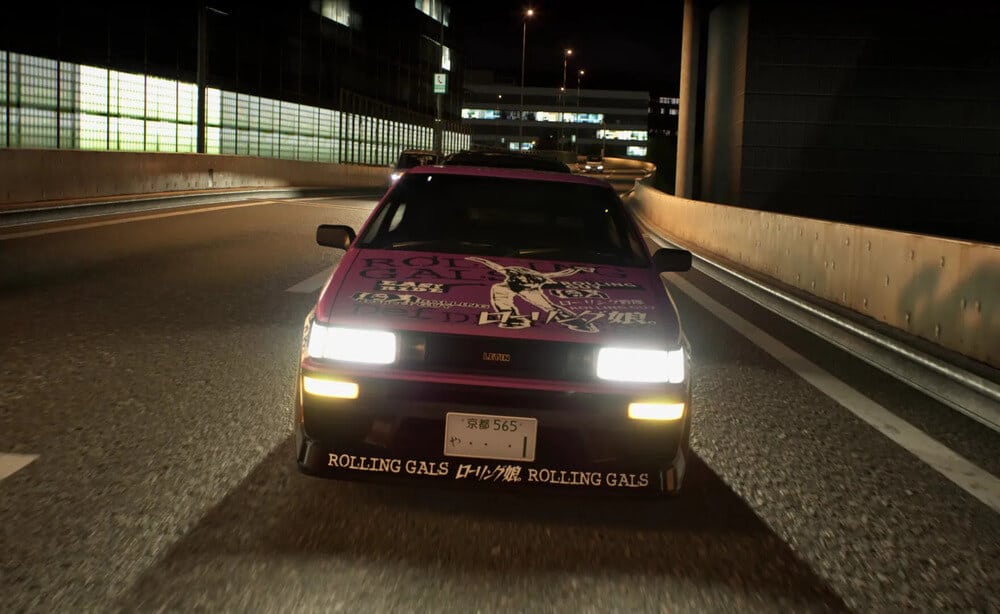 "La imagen muestra un coche conduciendo por una autopista de noche. El coche tiene un diseño distintivo con las palabras 'ROLLING GALS' destacadas en el capó y el parachoques delantero, junto con varios otros gráficos y textos en inglés y japonés. Los faros del coche están encendidos, iluminando la carretera. La matrícula es visible y dice 'や 565'. La escena parece ser de un videojuego, probablemente 'Tokyo Xtreme Racer', dado el contexto proporcionado.