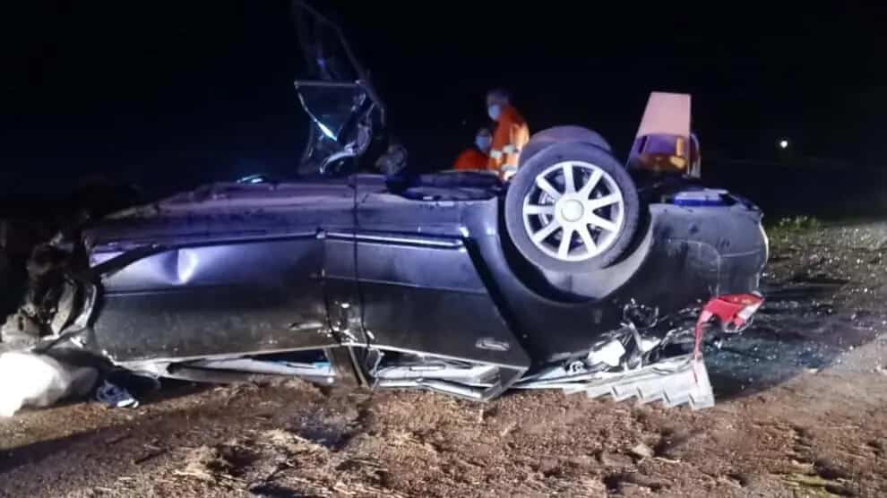 ALT text: "Imagen de un automóvil volcado completamente sobre su techo en lo que parece ser un accidente nocturno. El vehículo, de color oscuro, muestra daños significativos y está tumbado sobre un terreno irregular. Se puede ver una rueda con rin de aluminio expuesta y restos de la carrocería dañada. En el fondo se observa una luz naranja, posiblemente de servicios de emergencia.