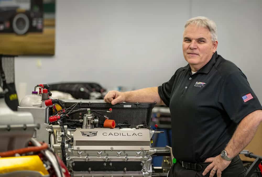 Russ O'Blenes, ingeniero de Cadillac, posando junto a un motor de competición con el logotipo de Cadillac en un taller técnico.