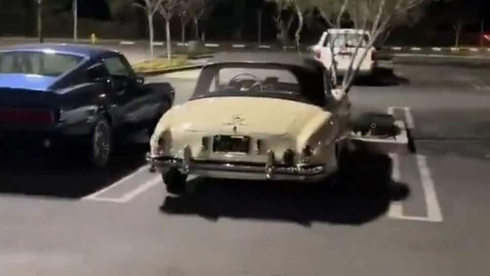Un aparcamiento nocturno con tres coches estacionados. El coche en el centro es un Mercedes-Benz 300SL Gullwing descapotable de color claro con la capota puesta, y parece ser un modelo antiguo. A la izquierda del descapotable hay un coche azul oscuro, y a la derecha, en el fondo, hay un coche blanco. La escena está iluminada por luces artificiales, y hay árboles sin hojas en el fondo. La imagen es interesante porque presenta un coche clásico, que podría ser de interés para los entusiastas o coleccionistas de automóviles.