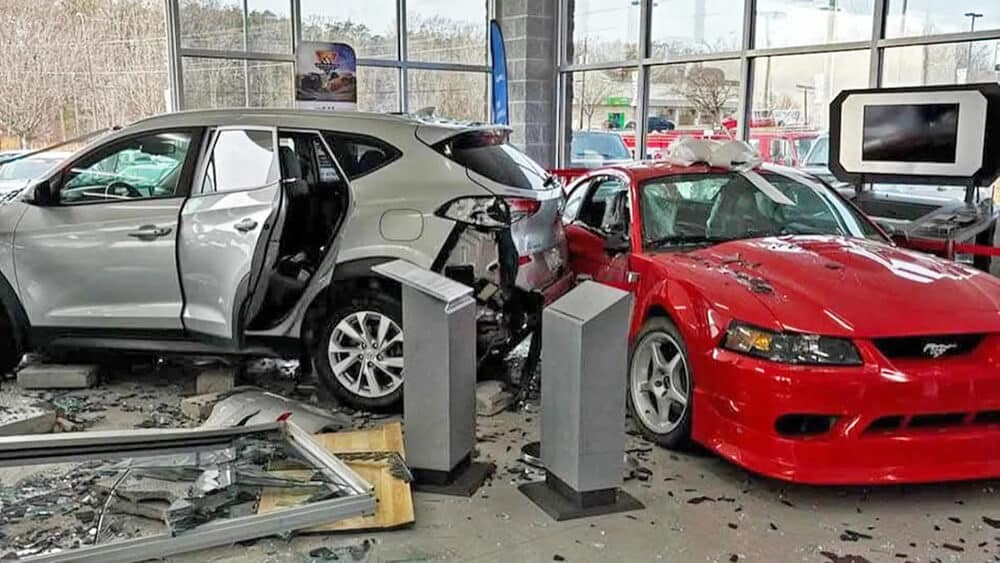 Imagen de un Ford Mustang SVT del año 2000 de color rojo, que ha sido impactado por un Hyundai Tucson. La Tucson ha atravesado el escaparate del concesionario, causando daños significativos en la parte trasera de la Tucson y en el lateral derecho del Mustang. No hay personas presentes en la escena.