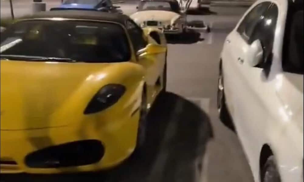 "Un Ferrari 430 amarillo está estacionado junto a un coche blanco en un aparcamiento. En el fondo, se puede ver un coche clásico, posiblemente antiguo. La imagen destaca la mezcla de vehículos modernos y clásicos en un solo cuadro, resaltando el contraste entre diferentes épocas del diseño automotriz.