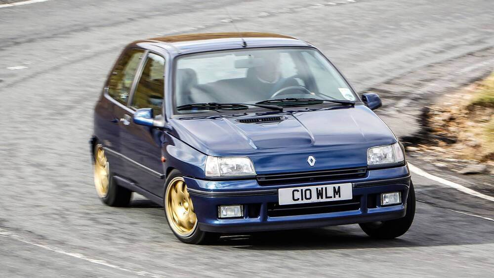 La imagen muestra un Renault Clio Williams azul, una versión de alto rendimiento del Renault Clio hatchback. El coche destaca por sus distintivas llantas de aleación doradas y su diseño deportivo. El vehículo está capturado en movimiento, probablemente en una carretera o pista, lo que resalta sus capacidades de rendimiento. La matrícula dice "C10 WLM", que podría ser una placa personalizada relacionada con el nombre del modelo "Williams".