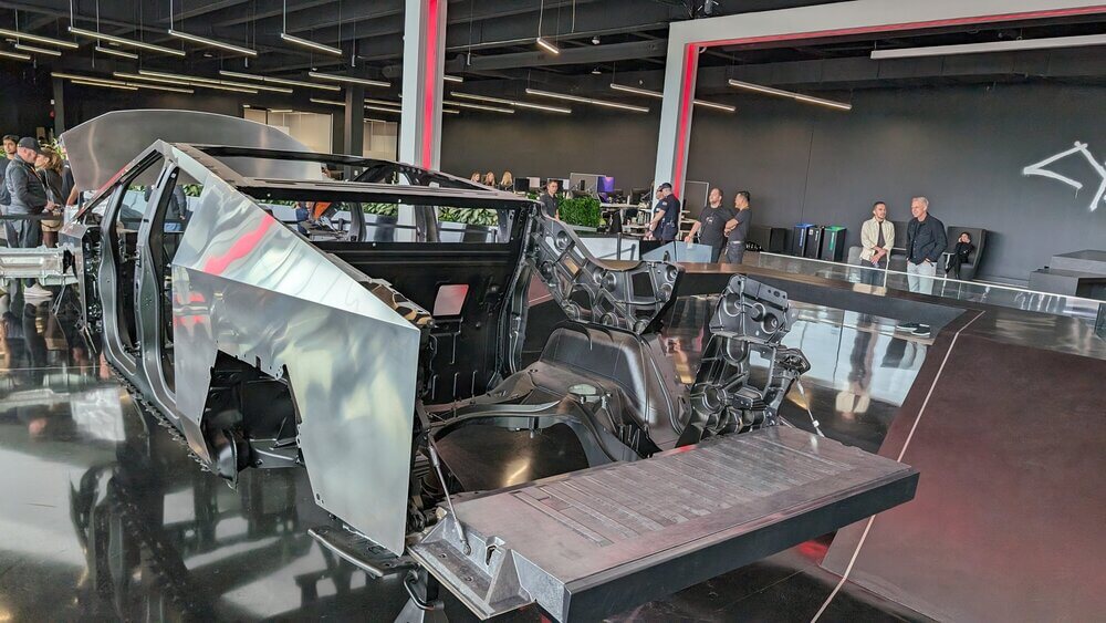 "Chasis de un vehículo, probablemente un prototipo de la Tesla Cybertruck, exhibido en un showroom o espacio de trabajo moderno e industrial. El chasis es metálico y está parcialmente ensamblado, con componentes estructurales visibles y un interior abierto. Varias personas están observando el chasis, lo que indica que podría tratarse de una demostración o un evento. El entorno incluye un suelo negro elegante y una iluminación moderna, lo que contribuye a una atmósfera de alta tecnología e innovación.