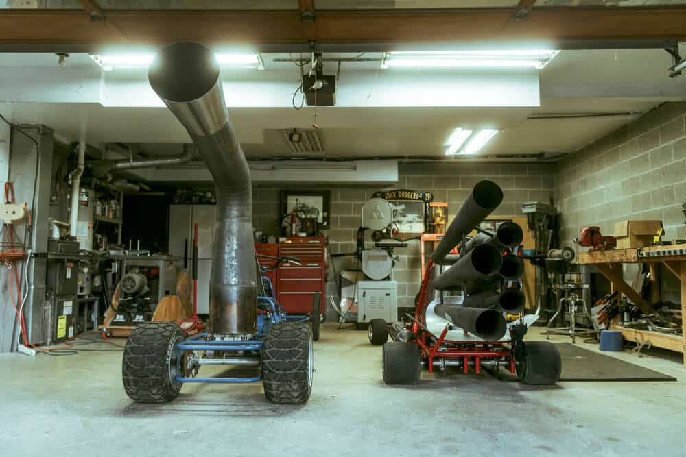 "El taller de Bob Maddox con dos vehículos a reacción caseros. El vehículo a la izquierda tiene un tubo de escape grande y curvado, mientras que el de la derecha tiene múltiples tubos de escape más pequeños. El taller está lleno de herramientas y equipos de trabajo, incluyendo una caja de herramientas roja, una sierra de mesa, y varias estanterías con suministros. La iluminación es proporcionada por luces fluorescentes en el techo.