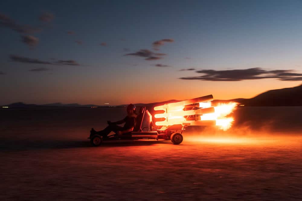 Fotografía dramática al atardecer de un go-kart equipado con motores pulse jet en funcionamiento. El vehículo está en movimiento sobre lo que parece ser un terreno desértico plano. Los motores pulse jet están encendidos, emitiendo llamaradas brillantes de color naranja intenso que contrastan espectacularmente con el cielo crepuscular. El conductor está sentado en posición de conducción, usando equipo de protección. El horizonte muestra montañas oscuras y un cielo con nubes dispersas iluminadas por los últimos rayos del sol. La escena está bañada en tonos naranjas y azules profundos, creando una imagen muy cinematográfica.