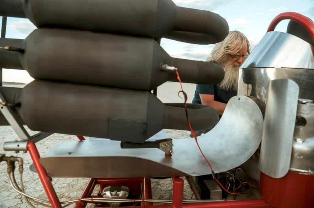 Imagen en primer plano de un motor pulse jet de gran tamaño montado sobre un vehículo rojo. El motor está pintado en color gris oscuro/negro mate y tiene una forma tubular característica con múltiples secciones. En el fondo se puede ver a una persona con barba larga y gris trabajando en el vehículo, vistiendo una camiseta azul oscuro. La imagen está tomada en un ambiente exterior con luz natural difusa.