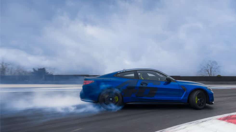 Un BMW M4 CS de color azul con el número "46" destacado en su lateral. El coche está derrapando en una pista de carreras, generando una cantidad significativa de humo de los neumáticos traseros. El fondo está desenfocado, lo que indica movimiento, y la escena parece estar ocurriendo en un día nublado.