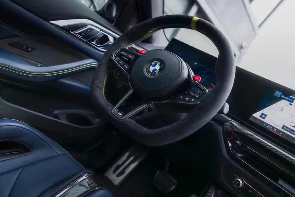 El interior de un coche BMW, específicamente enfocado en el volante y el área del tablero. El volante presenta el logotipo de BMW en el centro y tiene varios botones de control. El tablero incluye una pantalla digital que muestra un mapa y la información de navegación. El diseño interior incluye asientos de cuero azul con costuras amarillas y detalles en fibra de carbono, lo que indica un modelo de alto rendimiento, probablemente la edición BMW M4 CS VR46.