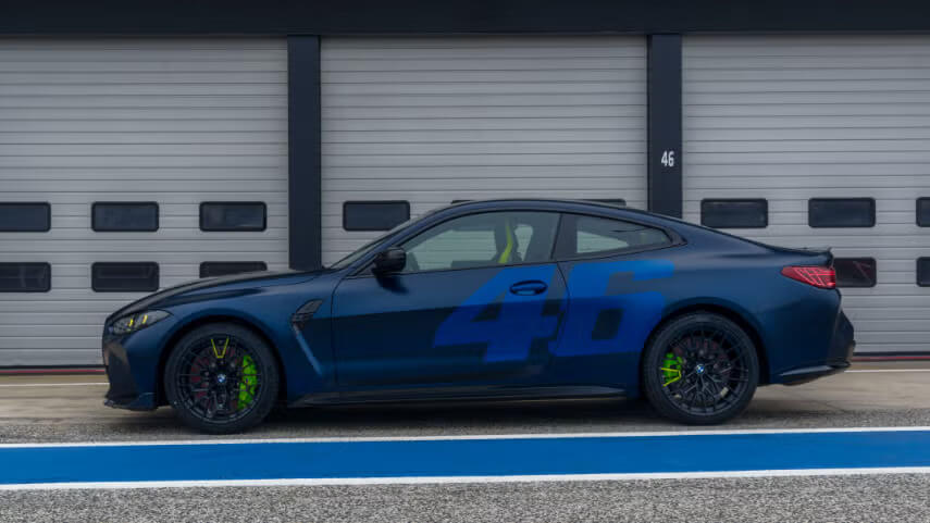 Vista lateral de un BMW M4 CS azul oscuro con el número "46" destacado en azul claro en el lateral. El coche está estacionado frente a un garaje con las puertas cerradas, y el número "46" también es visible en la puerta del garaje al fondo. El coche tiene llantas negras con pinzas de freno verdes, y el entorno parece ser una pista de carreras o una instalación similar.