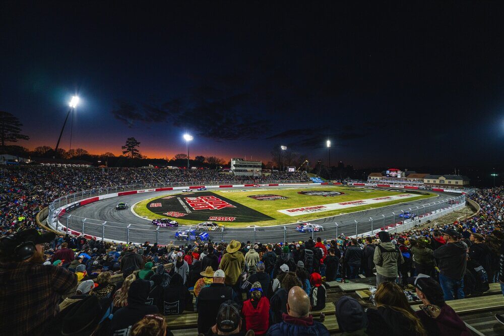 Imagen de una carrera nocturna en el Bowman Clash de NASCAR 2025. La pista es ovalada y está bien iluminada con múltiples focos. Varios coches de carreras están en movimiento en la pista. Las gradas están llenas de espectadores, lo que indica un alto nivel de interés y asistencia. El cielo está oscuro con un toque de luz del atardecer en el horizonte. La escena captura la emoción y energía de un evento en vivo de NASCAR.