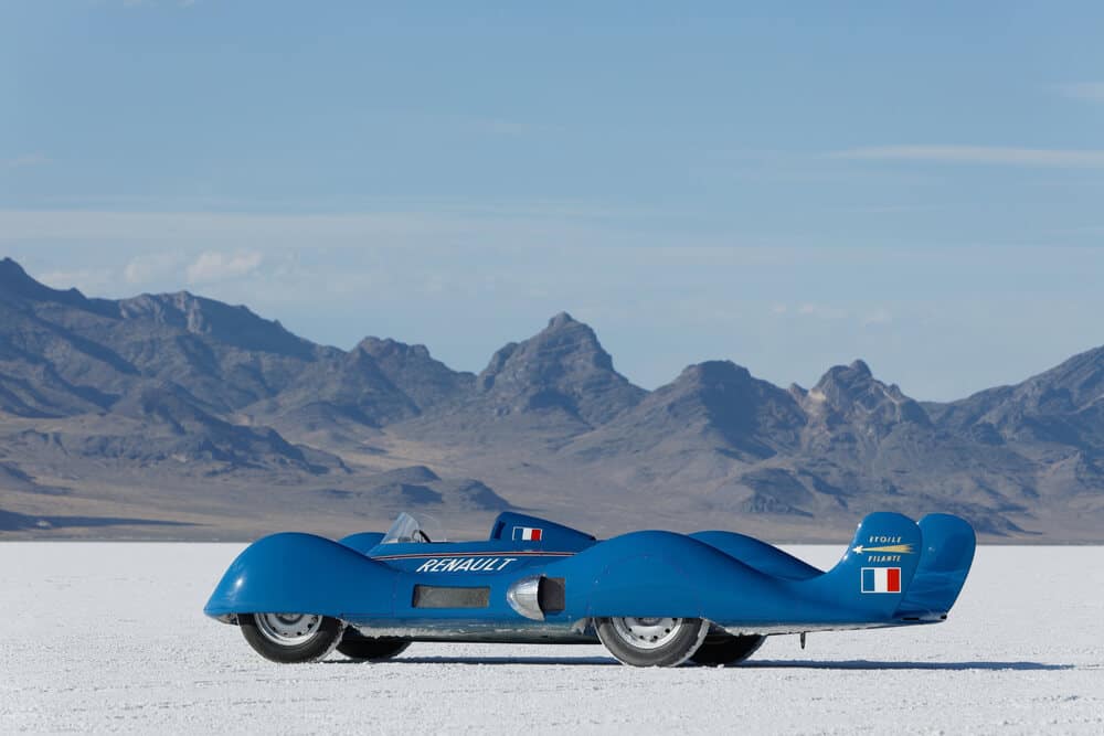 "El Renault Etoile Filante es un coche aerodinámico de color azul, situado en un paisaje de montañas y una superficie blanca y plana, probablemente las Salinas de Bonneville. El coche tiene un diseño elegante con ruedas cubiertas y un perfil bajo. El texto 'RENAULT' está destacado en el lateral del coche, junto con la bandera francesa. La parte trasera del coche también muestra el texto 'ETOILE FILANTE' y otra bandera francesa. Esta imagen es interesante y relevante porque el Renault Etoile Filante estableció un récord mundial de velocidad para coches con turbina en 1956 en las Salinas de Bonneville, alcanzando una velocidad de 306.9 km/h (190.9 mph).