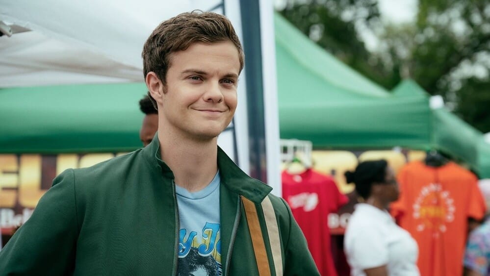 Jack Quaid caracterizado como Howie de The Boys, con una chaqueta verde y una camiseta azul con un diseño gráfico. En el fondo, hay puestos de mercado con camisetas colgadas y personas caminando.