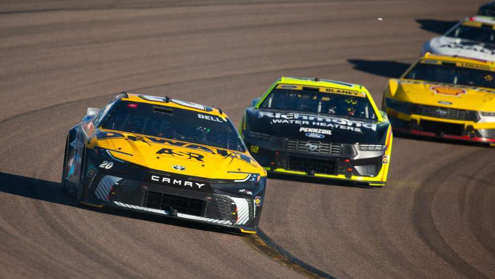 La imagen muestra un grupo de coches de carreras compitiendo en una pista de carreras. El coche en la delantera es un Toyota Camry amarillo con el número 20 y el nombre "BELL" en el parabrisas. El segundo coche es un Ford Mustang negro y amarillo con el número 12 y el nombre "BLANEY" en el parabrisas. Otros coches son visibles en el fondo, también compitiendo de cerca. Esta imagen es interesante y relevante porque captura la intensidad y competencia de una carrera profesional de coches, destacando la velocidad y habilidad involucradas en el automovilismo.