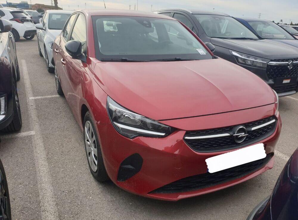 Una imagen del Opel Corsa 1.2 XEL Edition estacionado en una calle de Zaragoza. El coche es de color rojo brillante y tiene un diseño aerodinámico y moderno. En el fondo, se puede ver una vista urbana típica de Zaragoza, con edificios de varios pisos y árboles a lo largo de la calle. El Opel Corsa está en primer plano y ocupa la mayor parte de la imagen, mostrando su parte frontal y lateral.